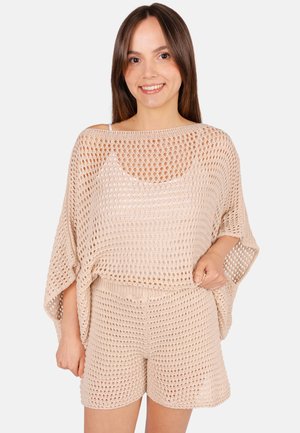 Jeune femme souriante portant un haut beige en maille ajourée et un short assorti par-dessus un débardeur blanc sur un fond uni.