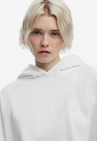 Hvid hættebluse med en blød tekstur, der har et broderet blomsterdesign på venstre side og en justerbar hætte.
