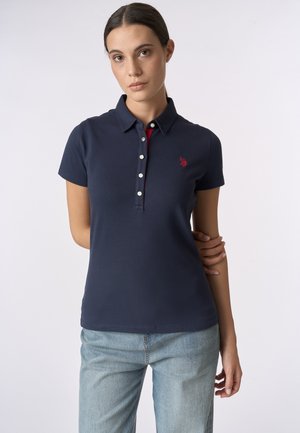 Giovane donna con polo blu navy a maniche corte con piccolo logo rosso e jeans azzurri chiari, in piedi davanti a uno sfondo semplice.