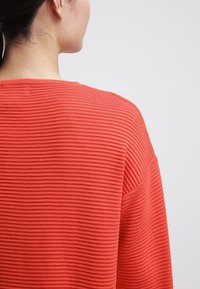 Röd ribbad tröja med en texturerad yta, avslappnad passform och vid halsringning, med subtila horisontella linjer över hela.