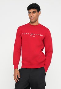 Felpa rossa con colletto tondo, caratterizzata da testo e logo bianchi "TOMMY HILFIGER EST. 1985". Realizzata in un tessuto strutturato, con polsini aderenti.
