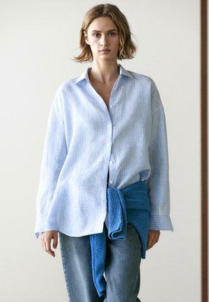 Jeune femme portant une chemise rayée bleu clair ample avec un pull bleu noué autour de la taille et un jean en denim.