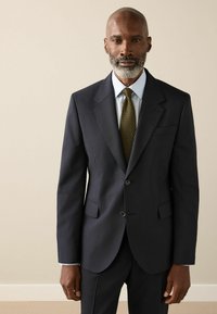 Dunkelblaues Sakko mit einreihigem Schnitt, Notch-Revers und zwei Knöpfen. Das Outfit wird durch ein hellblaues Hemd und eine olivgrüne Krawatte komplettiert.