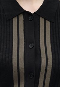 Polo en maille noir avec des rayures verticales beige et noires et trois boutons noirs au centre devant.