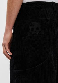 Fekete kordbársony nadrág, amelyen a hátsó zsebben egy feltűnő fekete koponya és keresztcsont hímzés található; függőleges bordázott textúrával.