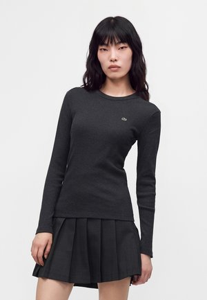 Vrouw met lang zwart haar, gekleed in een aansluitende donkere top met lange mouwen en een geplooide zwarte rok tegen een effen lichte achtergrond.
