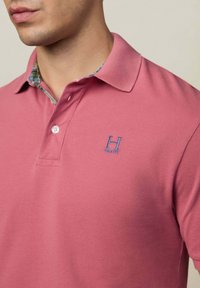 Ružový polo tričko vyrobené z textúrovanej tkaniny, s patterned golierom a malým modrým vyšitým logom na hrudi. Dva viditeľné gombíky.