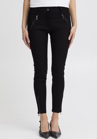 Žena u uskim crnim hlačama s patentnim zatvaračima sprijeda i na gležnjevima, uparenim s crnim cipelama s oštrim vrhom, stoji iza jednostavne pozadine.