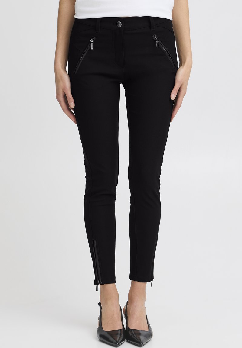 Žena u uskim crnim hlačama s patentnim zatvaračima sprijeda i na gležnjevima, uparenim s crnim cipelama s oštrim vrhom, stoji iza jednostavne pozadine.