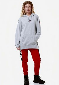 Felpa grigia con cappuccio, tasca frontale e logo, abbinata a pantaloni da jogging rossi con strisce nere e scritte bianche, indossata con stivali neri.