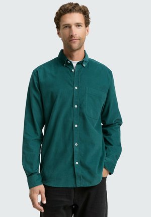 Chemise bleu canard à boutons avec une poche, confectionnée en tissu doux, dotée d'un col pointu et de manches longues, associée à un pantalon sombre.