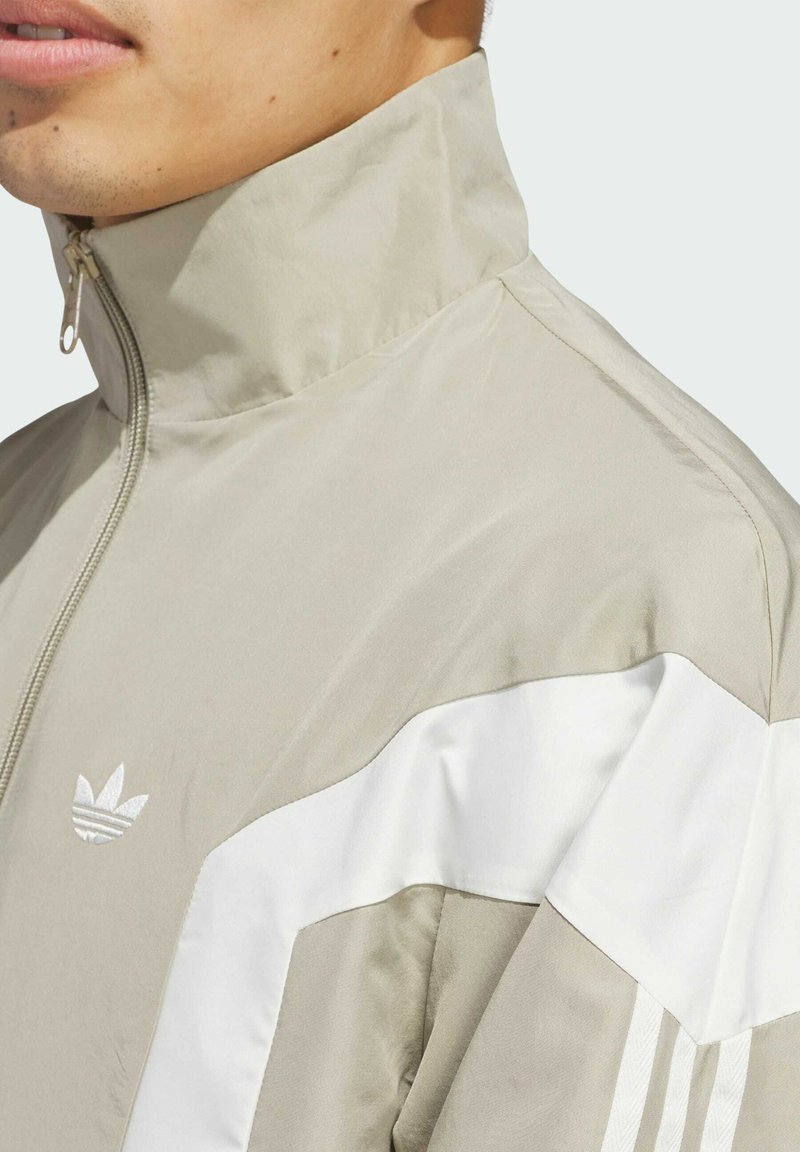 Light beige zip-up jacket with high collar, featuring white side panels and three white stripes. Soft, smooth texture, with logo on chest.