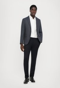 Dunkelblazer mit Muster über einem weißen Hemd, kombiniert mit slim-fit dunklen Hosen und schwarzen Schuhen. Glattes Material mit strukturiertem Design.