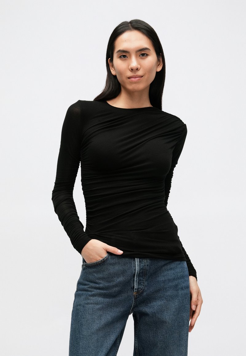 Sort langærmet top med tætsiddende design og tekstureret stof, der har rynkede detaljer langs torsoen. Bruges sammen med blå jeans.