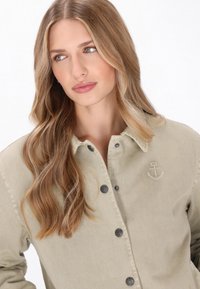 Khaki gombos ing tartós anyagból, finom horgonyhímzéssel és sötét gombokkal, laza szabással és klasszikus gallértervezéssel.