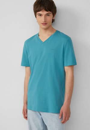 Turkusowa koszulka typu v-neck z miękkiej mieszanki bawełny, krótkie rękawy, z subtelnym detalem logo na piersi, w zestawie z jasnymi dżinsami.