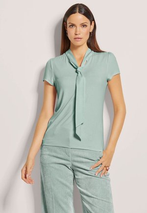 Vrouw die een lichtgroene korte mouwen blouse met neklintje en bijpassende corduroy broek draagt, staande tegen een effen lichte achtergrond.