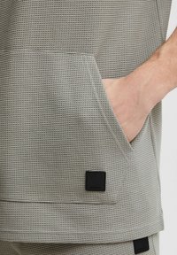Top gris texturizado con un patrón tejido, que presenta un gran bolsillo frontal y una etiqueta de marca negra en el dobladillo. Acabado de tela suave.