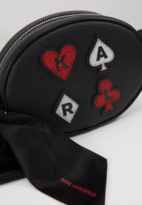 Svart läder oval crossbody-väska med färgglada applikationer, inklusive hjärtan och spader, samt ett svart satinband med röd text.