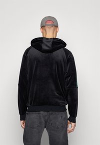 Felpa nera in velluto con cappuccio, dotata di maniche raglan e orlo curvo. Tessuto opaco, abbinata a jeans grigio scuro. Dettagli di design sottili.