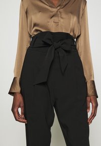 Femme portant un pantalon noir taille haute avec ceinture nouée en nœud et un chemisier marron brillant à manches longues avec larges poignets.
