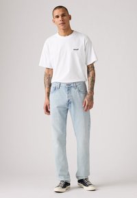 T-shirt bianca a maniche corte con logo nero di Levi's, abbinata a jeans azzurri chiari e sneakers nere basse. La pelle è tatuata su entrambe le braccia.