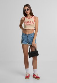 Zastrihnutý béžový top s červenými detailmi a textom "Von Dutch Originals", kombinovaný s roztrhnutými džínsovými šortkami s vyšitými detailmi a čiernou kabelkou.