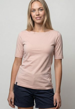 Giovane donna con lunghi capelli biondi che indossa una maglietta rosa chiaro a maniche corte e pantaloncini blu scuro con risvolto, in piedi davanti a uno sfondo semplice.