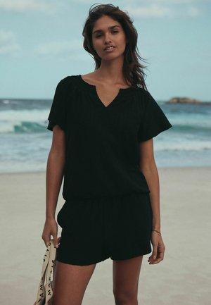 Fekete rövid ujjú felső V-nyakkal és húzott nyakkivágással, fekete rövidnadrággal párosítva. Az anyag könnyűnek és texturáltnak tűnik.