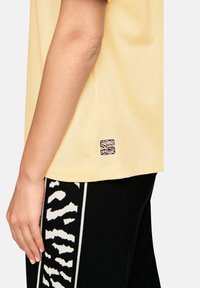 Yellow cotton t-shirt with short sleeves, featuring a small black-and-white logo on the side. Paired with black pants with a white abstract pattern.