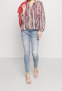 Mujer con una blusa a rayas colorida, jeans ajustados desgastados, tacones blancos con tiras y sosteniendo una chaqueta roja sobre su hombro.