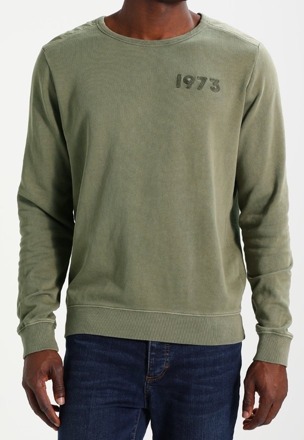Grön sweatshirt med lång ärm och ribbat rundhals, med året "1973" broderat i mörkgrön färg på bröstet, ihop med denimjeans.