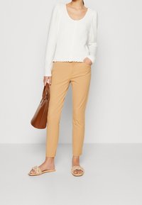 Witte textuur lange mouwen top, beige aangesloten broek, bruine handtas en lichtbeige slide sandalen met decoratieve knoopdetails.