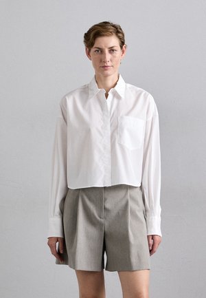 Woman with short hair wearing a white long-sleeve cropped shirt and high-waisted gray shorts standing against a plain light wall.