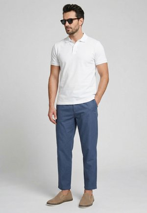 Mand iført sorte solbriller, hvid kortærmet polotrøje, blå bukser og beige slip-on sko, står med den ene hånd i lommen.