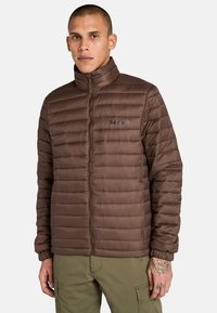 Brun puffjacka med kviltat mönster, hög krage och dragkedjestängning. Har elastiska ärmslut och Timberland-logotyp på bröstet.