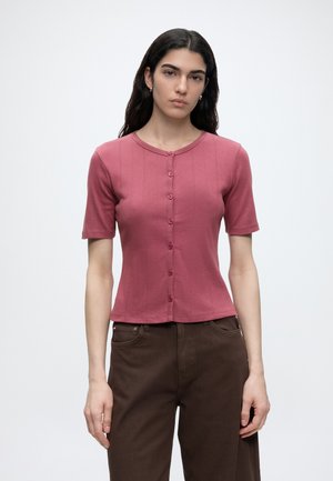 Femme aux longs cheveux foncés portant un haut rose à manches courtes avec boutons et un pantalon marron foncé, debout devant un fond clair uni.