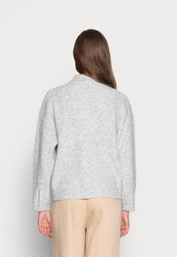 Hellgrau gestrickter Pullover mit lässiger Passform, geripptem Muster und steilem Kragen, kombiniert mit beigen Hosen. Details umfassen lange Ärmel.