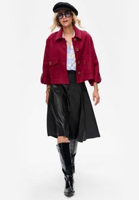 Carmijn geknipte jacket met grote knopen over een lichtblauw gemusterd shirt; zwarte leren midi rok en zwarte laarzen tot de knie.