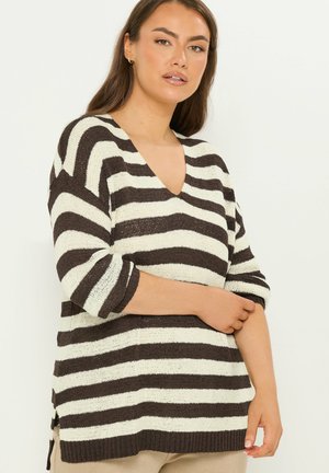 Kvinde iført en sort og hvid vidstribet V-hals sweater med trekvartlange ærmer, stående mod en ensfarvet lys baggrund.