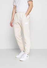 Man i off-white sweatpants med fickor och vita Adidas-sneakers, stående mot en enkel ljus bakgrund.