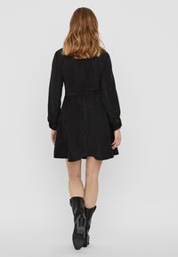 Schwarzes Wildlederkleid mit langen Ärmeln, taillierter Taille, weit schwingendem Rock und einem Gürtel. Kombiniert mit kniehohen schwarzen Stiefeln.
