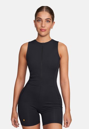 Frau mit zurückgekämmtem Haar, die einen ärmellosen schwarzen Sport-Overall mit Reißverschluss vorne vor einem schlichten weißen Hintergrund trägt.