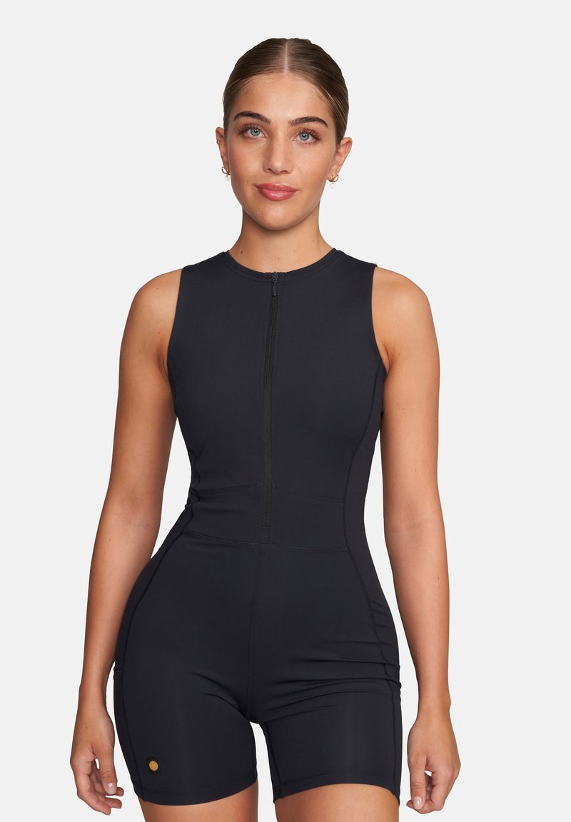 Kobieta z zaczesanymi do tyłu włosami w czarnym sportowym kombinezonie bez rękawów z zamkiem z przodu na jednolitym białym tle.