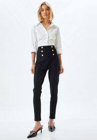 Witte knoopshirts gecombineerd met hoog uitgesneden zwarte broek met gouden knopen, op maat gemaakt, en glanzende zwarte hoge hakken.