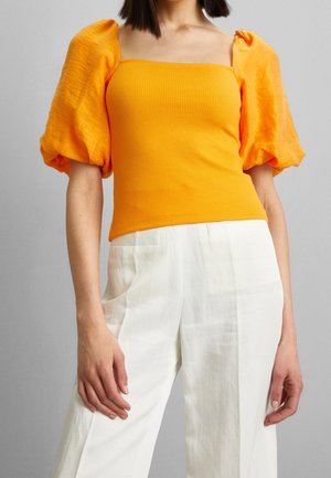 Vrouw draagt een feloranje top met pofmouwen en een vierkante halslijn en witte, hooggesneden wijde broek.
