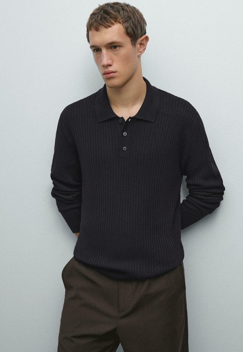 Jonge man in een zwart geribbeld langarm polo-shirt en donkerbruine broek, staand tegen een effen lichtgrijze muur.