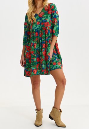 Kobieta w kwiecistej sukience do kolan w kolorach zielonym, niebieskim i czerwonym, z rękawami o długości do połowy przedramienia, w beżowych botkach, stoi z jedną nogą lekko wysuniętą do przodu.