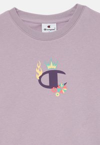 Liliowa bluza z fioletowym logo "C" z koroną, płomieniami oraz floralnymi akcentami w kolorach żółtym, zielonym i różowym.