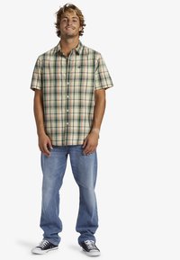 Camisa de cuadros de manga corta en beige, verde y naranja, con cuello abotonado y bolsillo en el pecho, combinada con jeans azul claro y zapatillas negras.